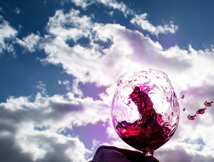 Una copa de vino tinto está en el aire con unas nubes un poco rojas y unas gotas de vino se caen de la copa, ya que el vino esta en movimiento.