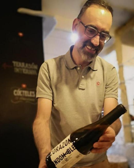 Manuel de Bodegas Mazuela, sonriente, sosteniendo una botella de vino en un ambiente acogedor de bodega.