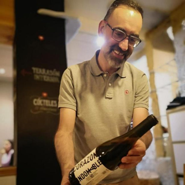 Manuel de Bodegas Mazuela, sonriente, sosteniendo una botella de vino en un ambiente acogedor de bodega.