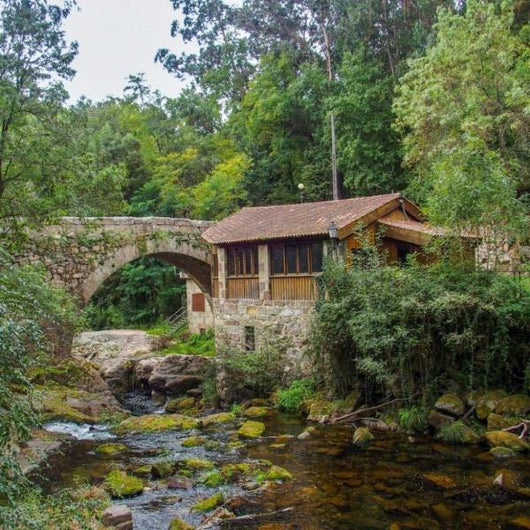 Puente de piedra sobre un arroyo con una casa rústica rodeada de vegetación exuberante en Arbo, cerca de bodegas Veigamoura.