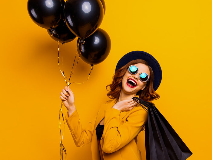 Chica sonriendo con gafas y un traje amarillo, muy alegre y con unos globos y una bolsa de la compra negra, montaje de Black Friday.