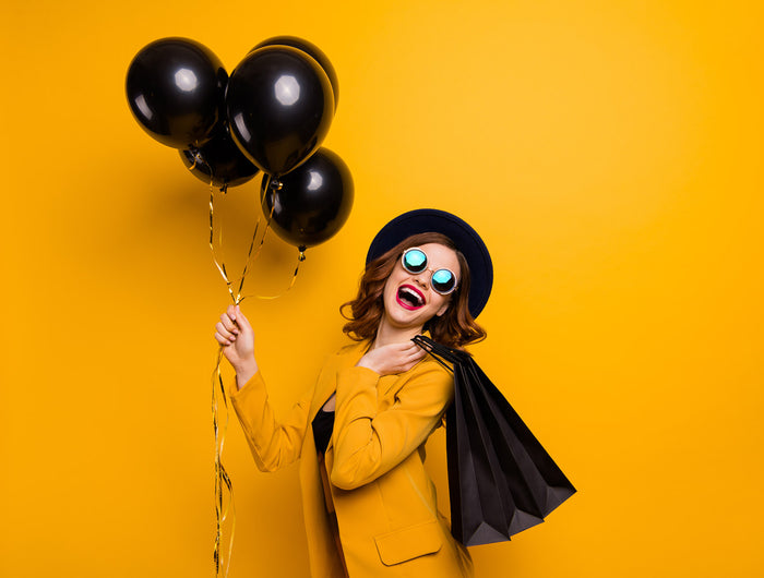 Chica sonriendo con gafas y un traje amarillo, muy alegre y con unos globos y una bolsa de la compra negra, montaje de Black Friday. Versión PC