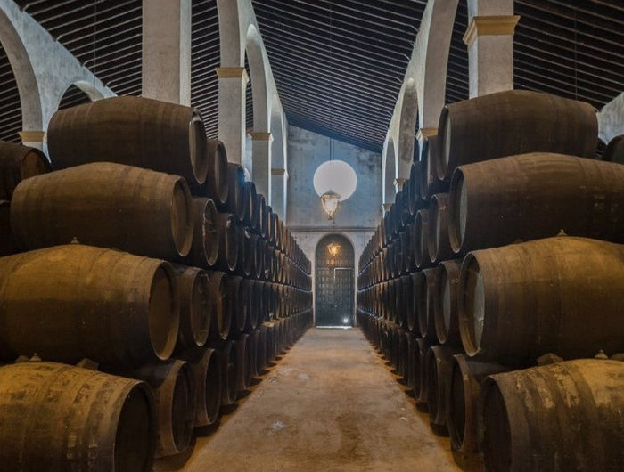 Una bodega típica del Marco de Jerez, con techos altos y columnas árabes, con una puerta al fondo y barriles de vino, a los lados del pasillo, apilados en 4 alturas.