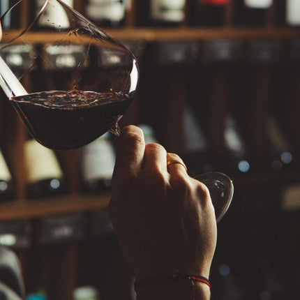 Persona catando un vino tinto en una copa, con un fondo de estantes de vino en una tienda de vinos iluminada tenue.