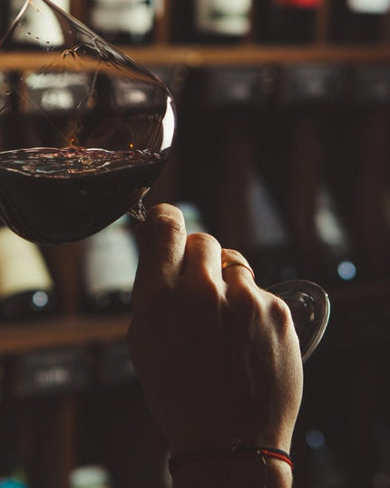 Persona catando un vino tinto en una copa, con un fondo de estantes de vino en una tienda de vinos iluminada tenue.